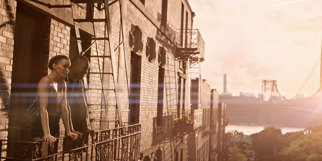 Image from In The Heights movie. Leslie Grace and Corey Hawkins standing on a fire escape with the GWB in the background.